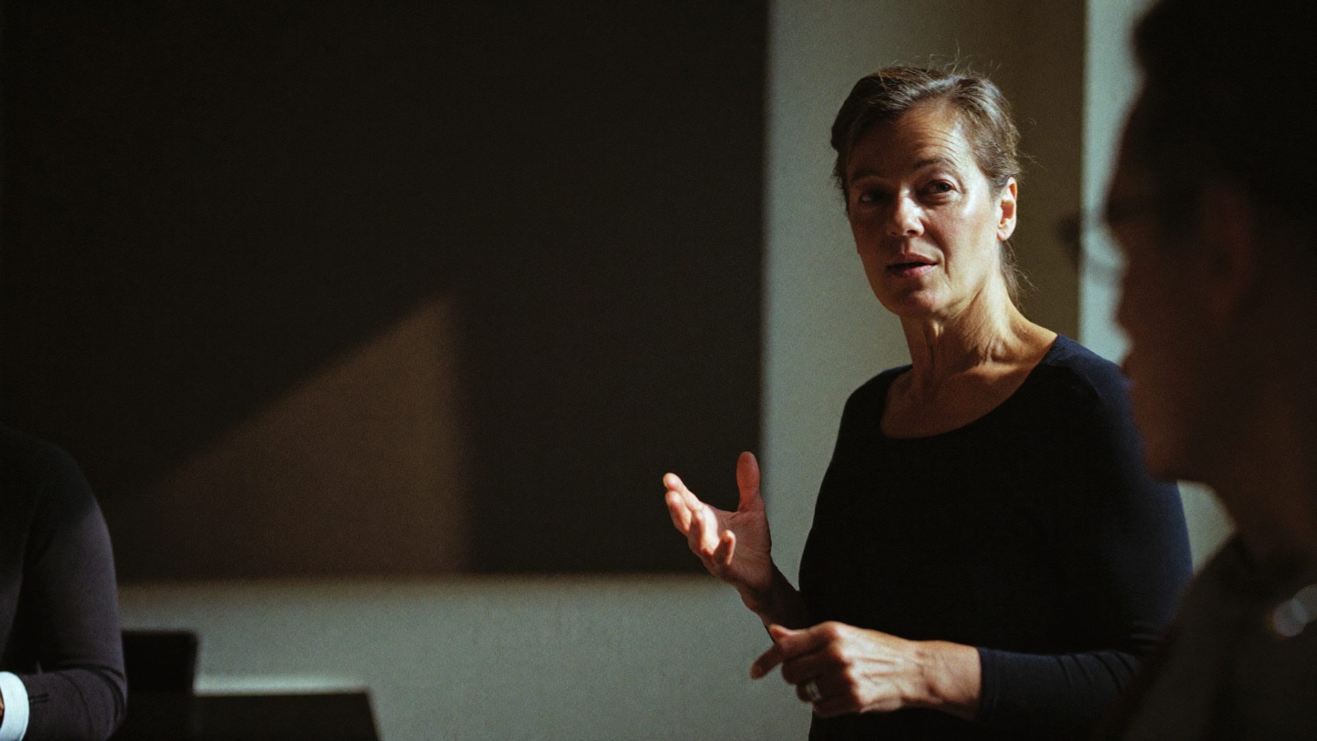 A teacher speaking to a small class, one hand raised as she explains a point to the people at the table
